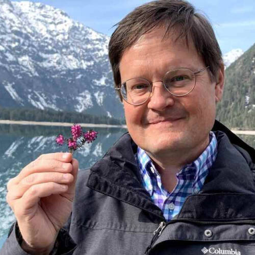 A man wearing glasses and a dark Columbia jacket smiles while holding a small purple flower in front of a scenic lake surrounded by snow-capped mountains and pine trees. The clear blue sky and reflections on the water suggest a calm, cool day in a mountai